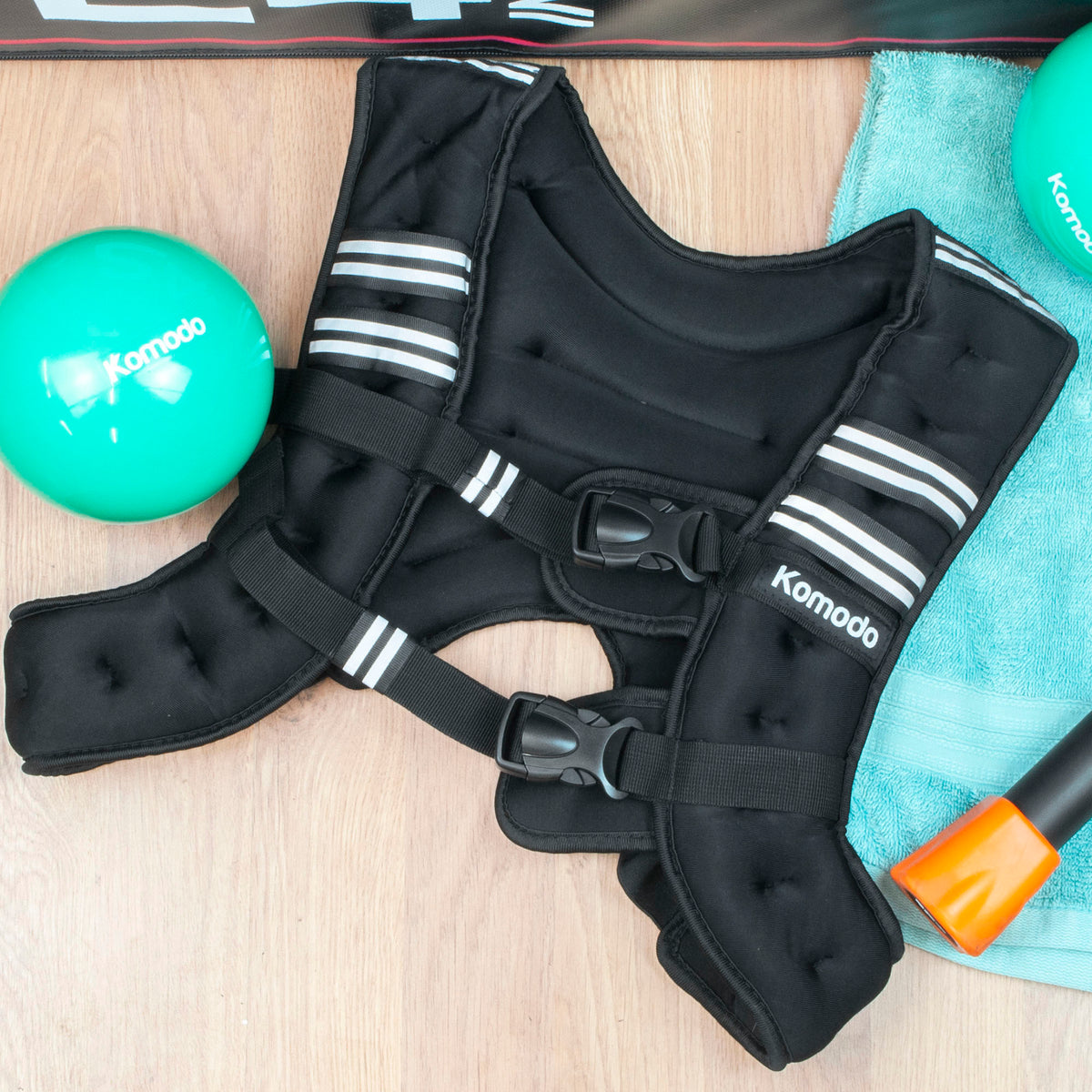A black Komodo weight vest with white stripes and black straps, laid flat on a light wood surface with a teal towel, a green ball, and part of another green ball visible.