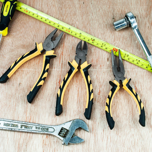Three pairs of hand tools on a light wood surface: two yellow-and-black pliers and one yellow-and-black wirenut pliers, with a yellow tape measure and a silver wrench above them.