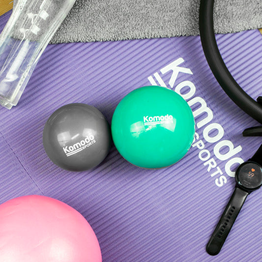 Two Komodo Sports exercise balls, one gray and one mint green, placed on a purple Komodo Sports mat alongside fitness accessories including a pink ball, smartwatch, and a clear water bottle.