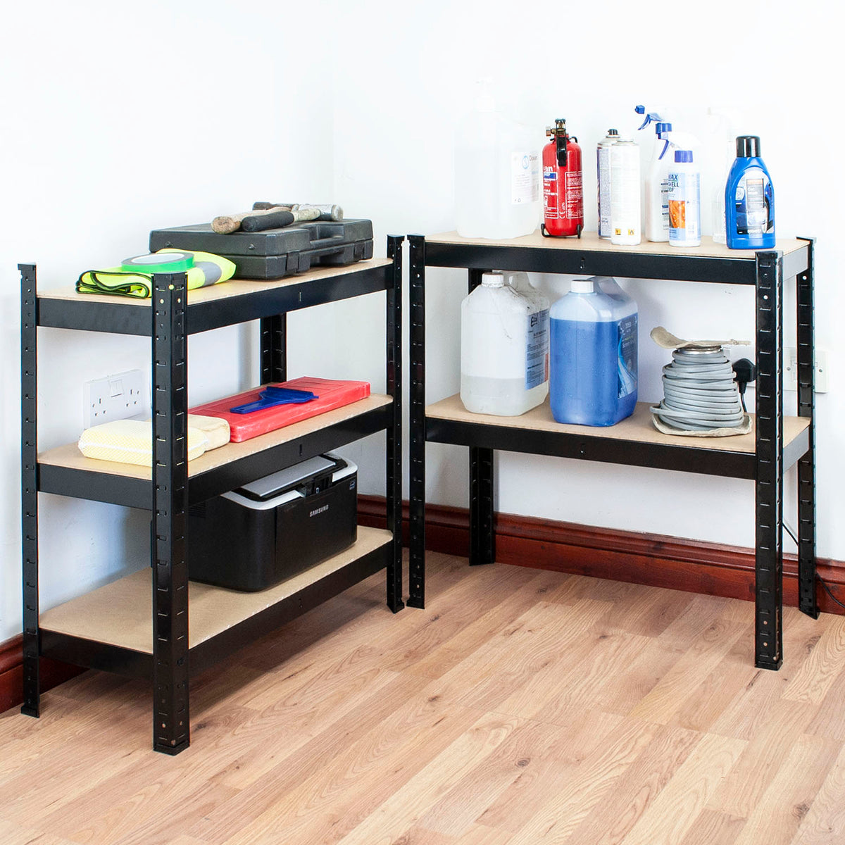 Two black metal industrial shelving units with light wood shelves, shown in a room corner with wood flooring. The shelves are stocked with various household and utility items including bottles, a fire extinguisher, and storage boxes.