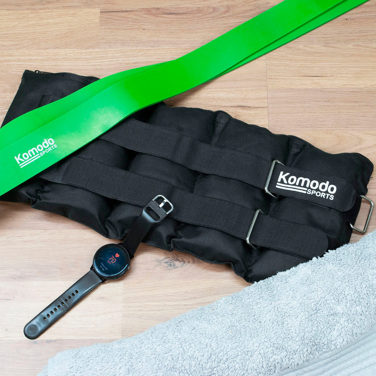 A black Komodo Sports ankle weight laid flat on a light wood floor, with a bright green resistance band and a black smartwatch placed on top. A light gray towel is visible in the lower right corner.