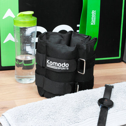 A black Komodo Sports ankle weight shown on a wooden floor with a water bottle, towel, and smartwatch nearby. The product is the ankle weight, photographed in a styled setup with branded packaging in the background.