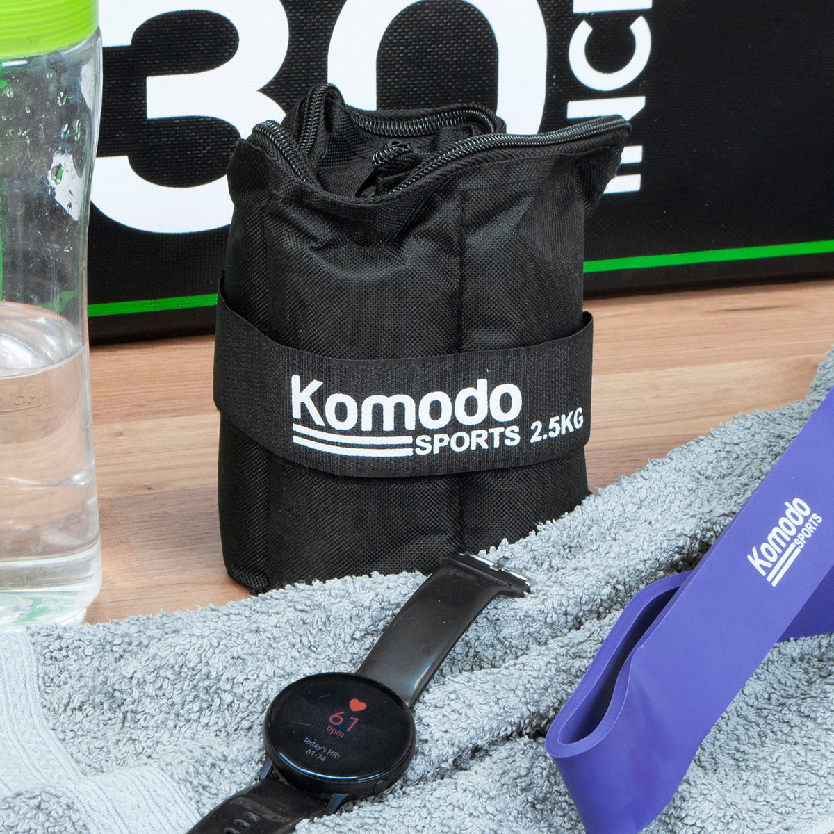 A black ankle weight with a white printed 'Komodo Sports 2.5KG' logo, placed on a wooden surface with a grey towel, smartwatch, purple resistance band, and water bottle nearby.
