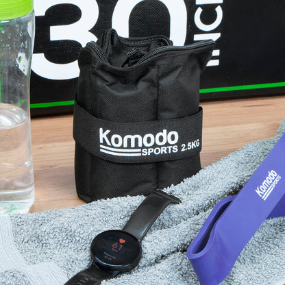 A black ankle weight with a white printed 'Komodo Sports 2.5KG' logo, placed on a wooden surface with a grey towel, smartwatch, purple resistance band, and water bottle nearby.