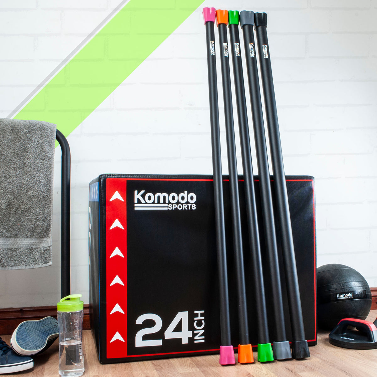 Komodo Sports weight bars. standing upright next to a Komodo Plyo box, on a wooden floor, with a towel, water bottle, and exercise ball in the background.