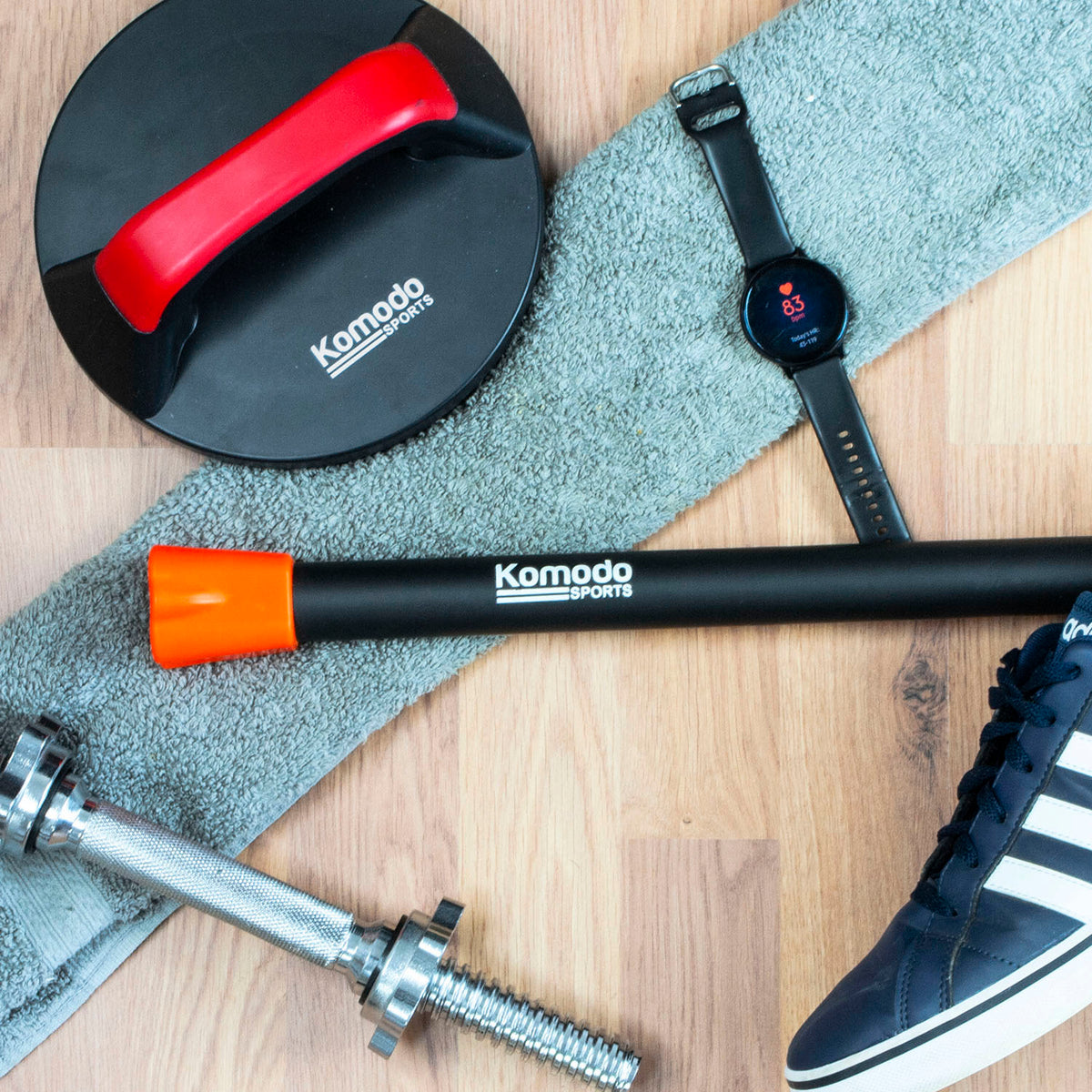 A a 4kg weighted bar Komado Sports set on a gray towel over a wooden floor, alongside a smartwatch, push handle and a trainer.