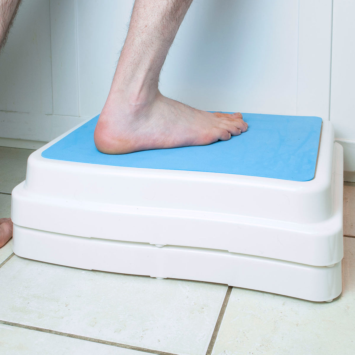 A white plastic bath step platform with a bright blue non-slip top surface, shown with a person's bare foot stepping onto it in a tiled indoor setting.
