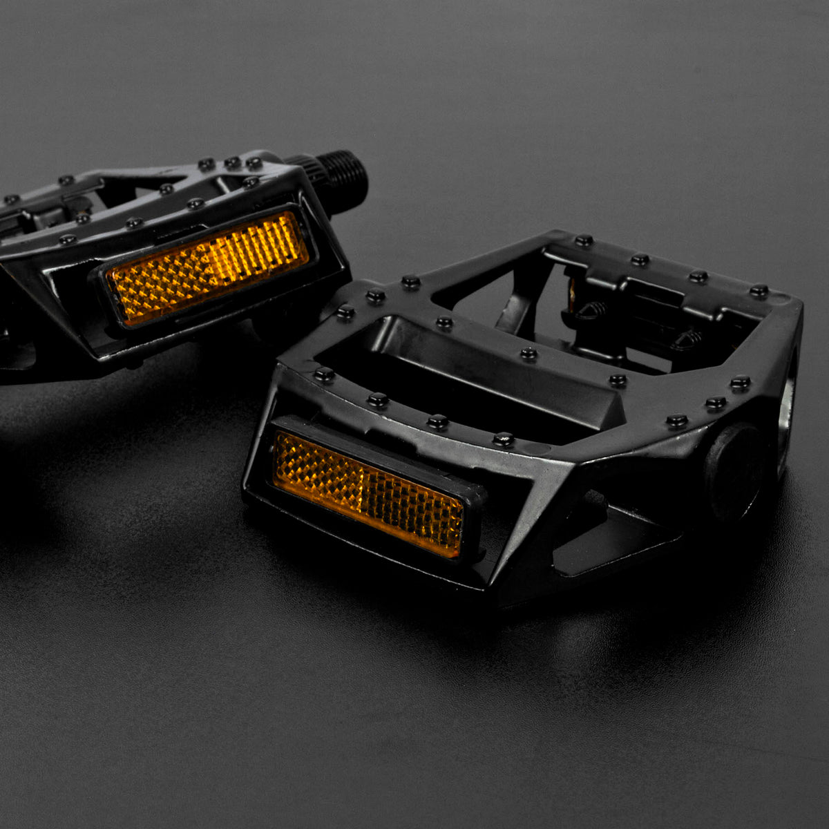 A pair of black metal bicycle pedals with integrated amber reflectors, photographed on a dark gray studio surface.