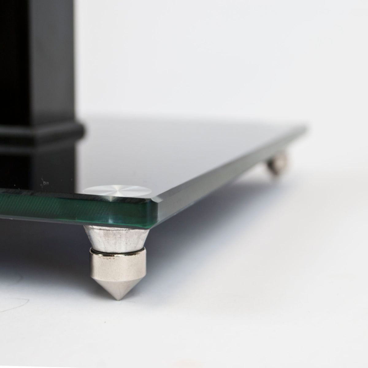 Close-up of a black glass speaker stand with silver metal cone feet and a black support leg, on a white background.