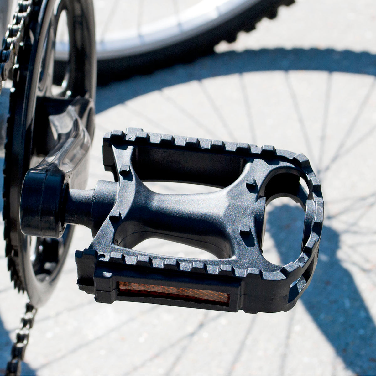 A close-up of a black bicycle pedal with a textured treaded edge, attached to a crankset, photographed outdoors on pavement with the bicycle wheel and shadow visible in the background.