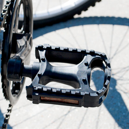 A close-up of a black bicycle pedal with a textured treaded edge, attached to a crankset, photographed outdoors on pavement with the bicycle wheel and shadow visible in the background.