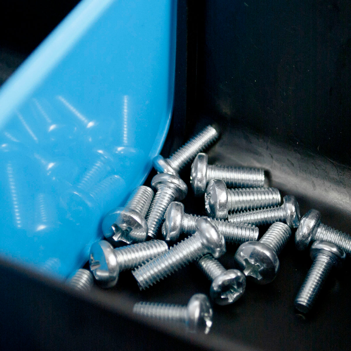 A pile of silver metal Flathead screws with cross-drive heads scattered inside a black container next to a bright blue plastic divider.