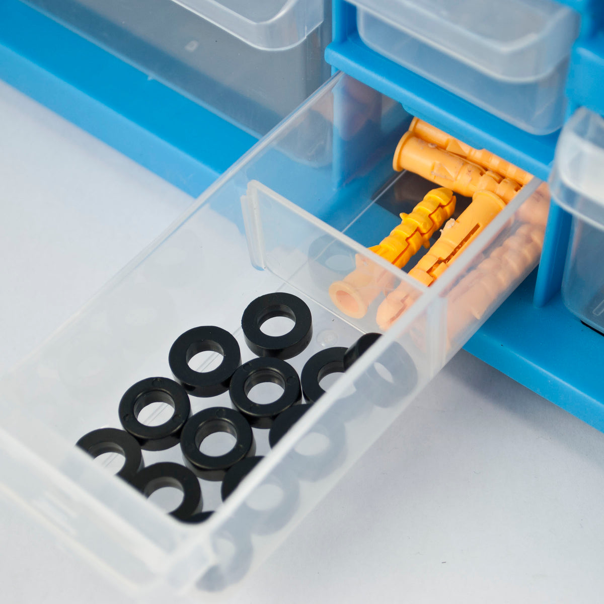 A close-up of a clear plastic drawer organizer with a pulled-out compartment holding black rubber grommets and yellow wall plugs inside a blue storage box.
