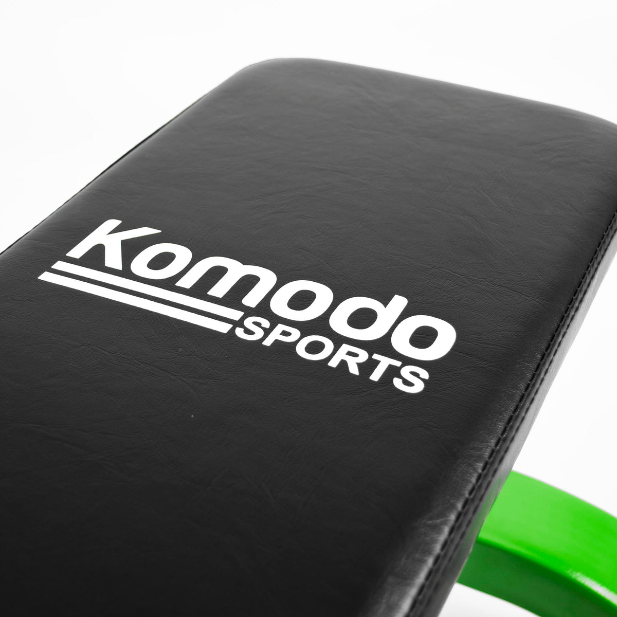 Close-up of a black padded fitness bench or weight bench with white Komodo Sports branding on the backrest, with a bright green frame edge visible at the bottom right against a white background.