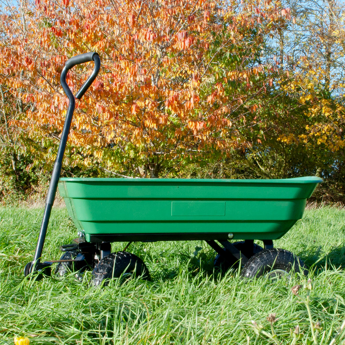 A green plastic garden dump trolley with black wheels and a black D-handle, shown in a grassy outdoor setting with autumn trees in the background.