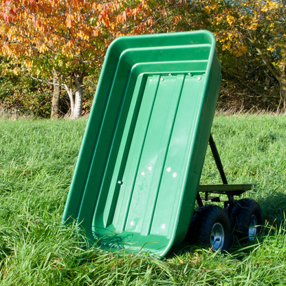 A green plastic dump trolley with a slanted scoop and black wheels, photographed outdoors in a grassy field with autumn trees in the background.