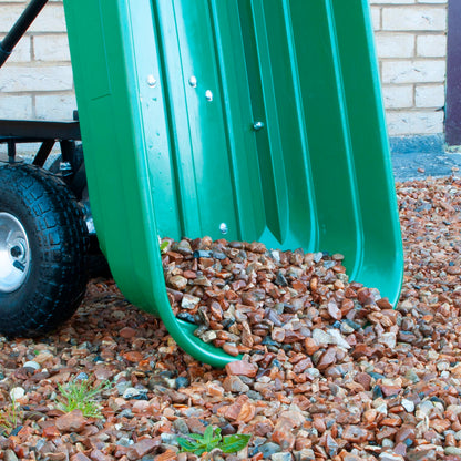 A green plastic dump trolley bucket filled with reddish-brown gravel, shown outdoors with a brick wall and ground covered in stones.