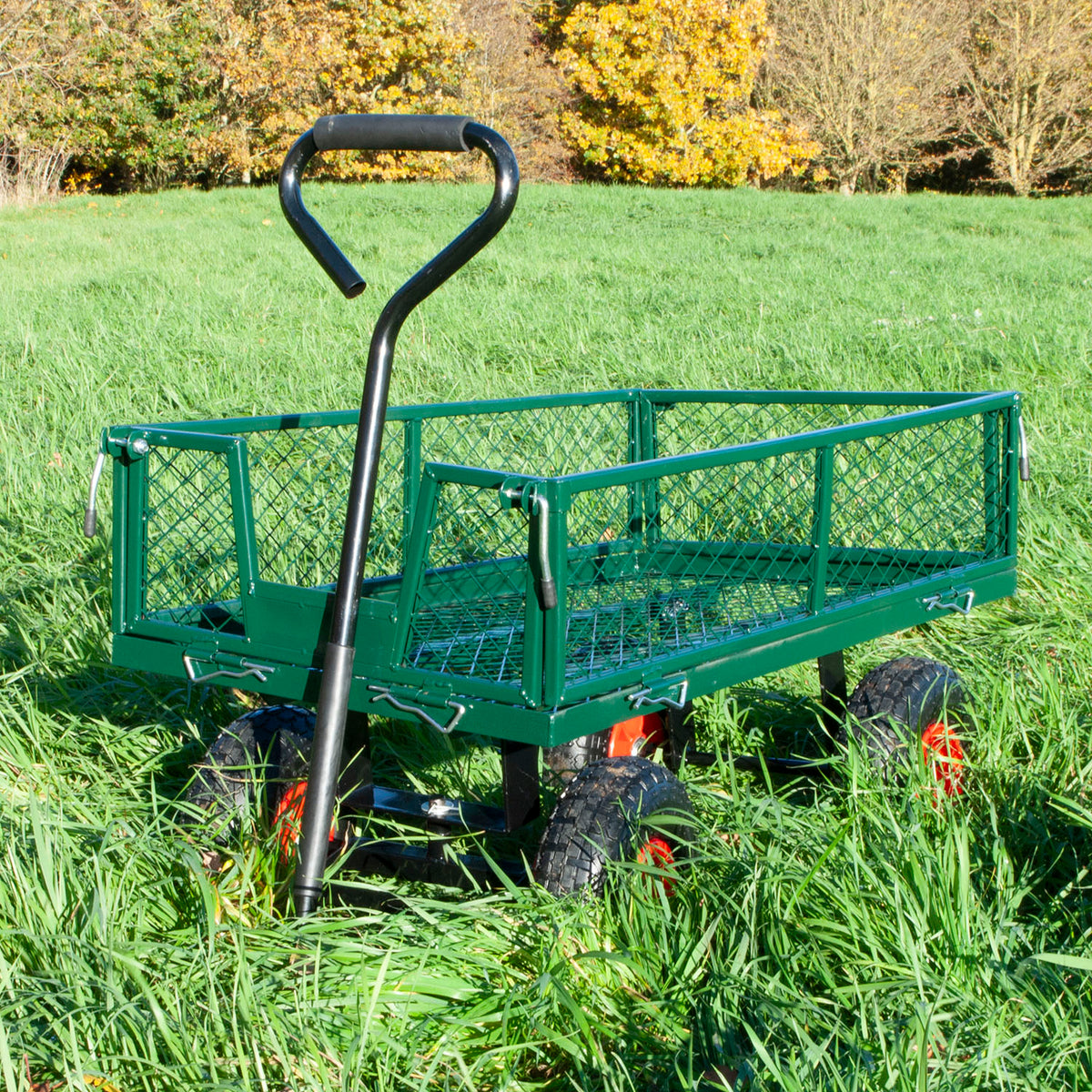 A green metal garden wagon with a black handle and mesh sides, shown in tall grass.
