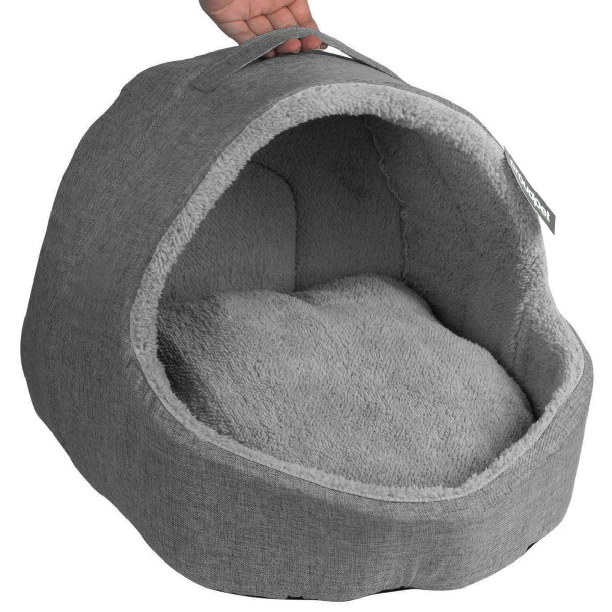 A gray fabric-covered pet bed with a plush gray cushion inside and a handle on top, shown against a white background. A human hand is visible holding the handle, which classifies the subject_type as Lifestyle.