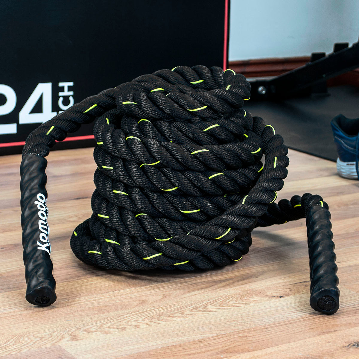 A coiled black battle rope with bright neon yellow-green stitching and textured black handles, resting on a light wood floor in a gym setting. The handle on the left is labeled 'Komodo'.