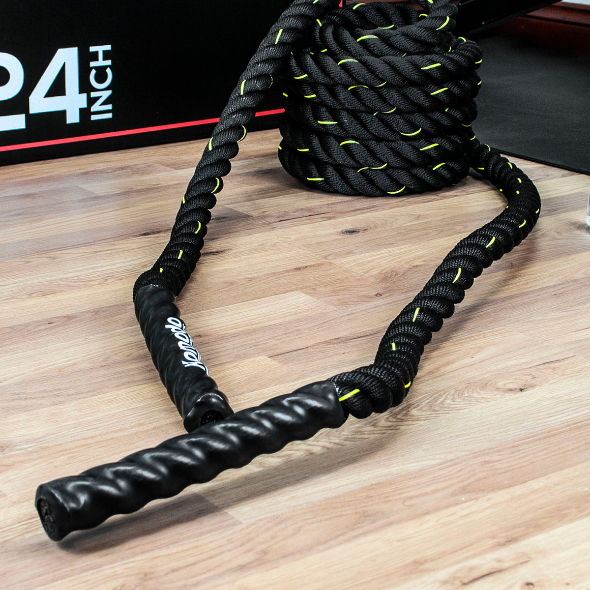 Black battle ropes with bright yellow-green stitching and a twisted handle, shown on a wooden floor with a coiled roll of the same ropes behind it.
