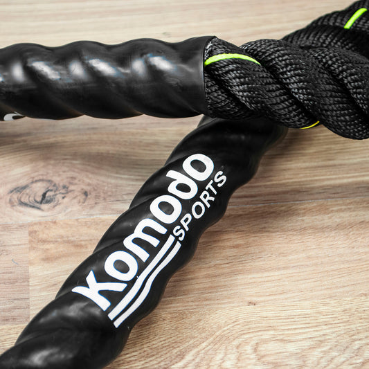 Black Komodo Sports battle ropes with white logo text and neon green accent stitching, shown on a light wood surface.