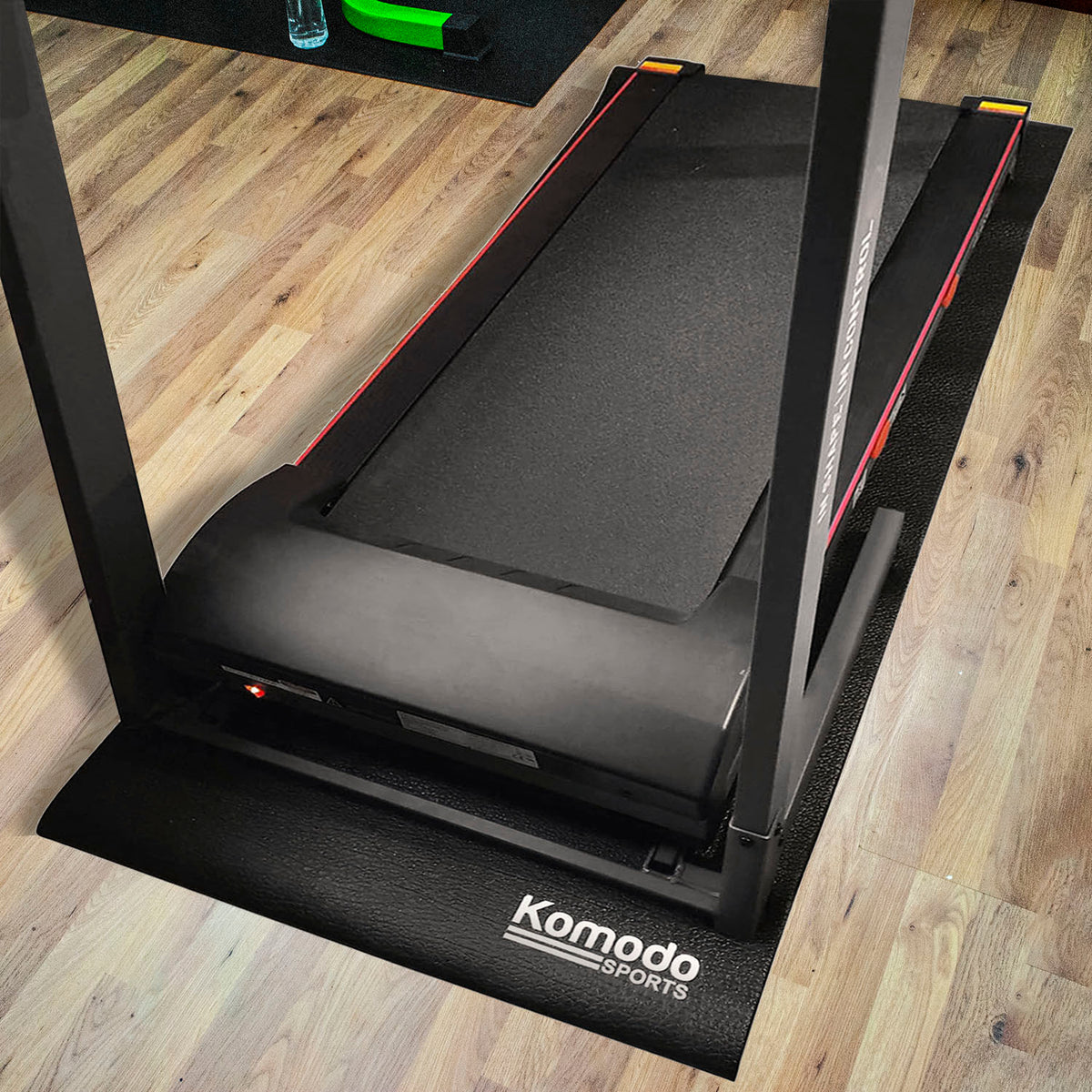 A black Komodo Sports treadmill, bike and gym mat placed under a treadmill on a light wood floor. The mat features the white Komodo Sports logo on the front corner.