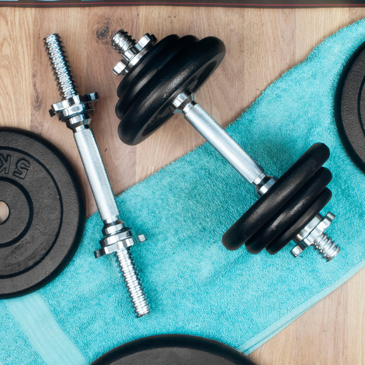Two adjustable dumbbells with black weight plates and silver metal rods, arranged on a turquoise towel over a light wood surface with additional black weight plates partially visible.