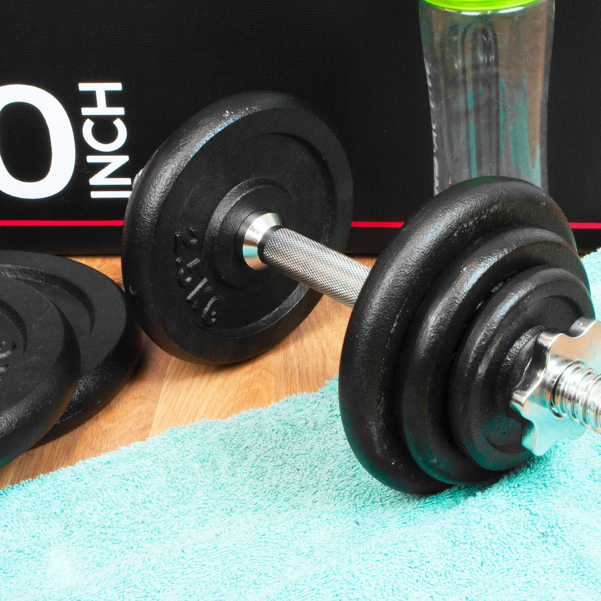 A close-up of a black adjustable dumbbell with a silver metal handle resting on a mint green towel, with a black box and a clear bottle in the background.
