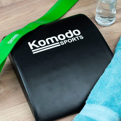 A black foam ab pad mat with white 'Komodo Sports' branding, shown on a wooden tabletop with a green resistance band, a clear water bottle, and a light blue towel.
