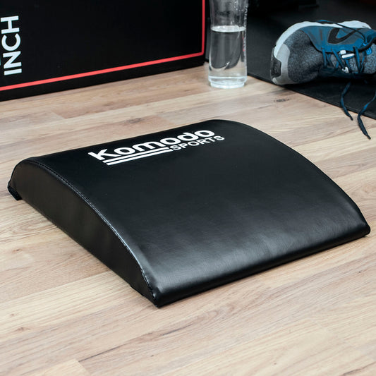A black curved foam roller pad with white 'Komodo Sports' branding, placed on a light wood floor with a shoe, water bottle, and part of a plyo box visible in the background.