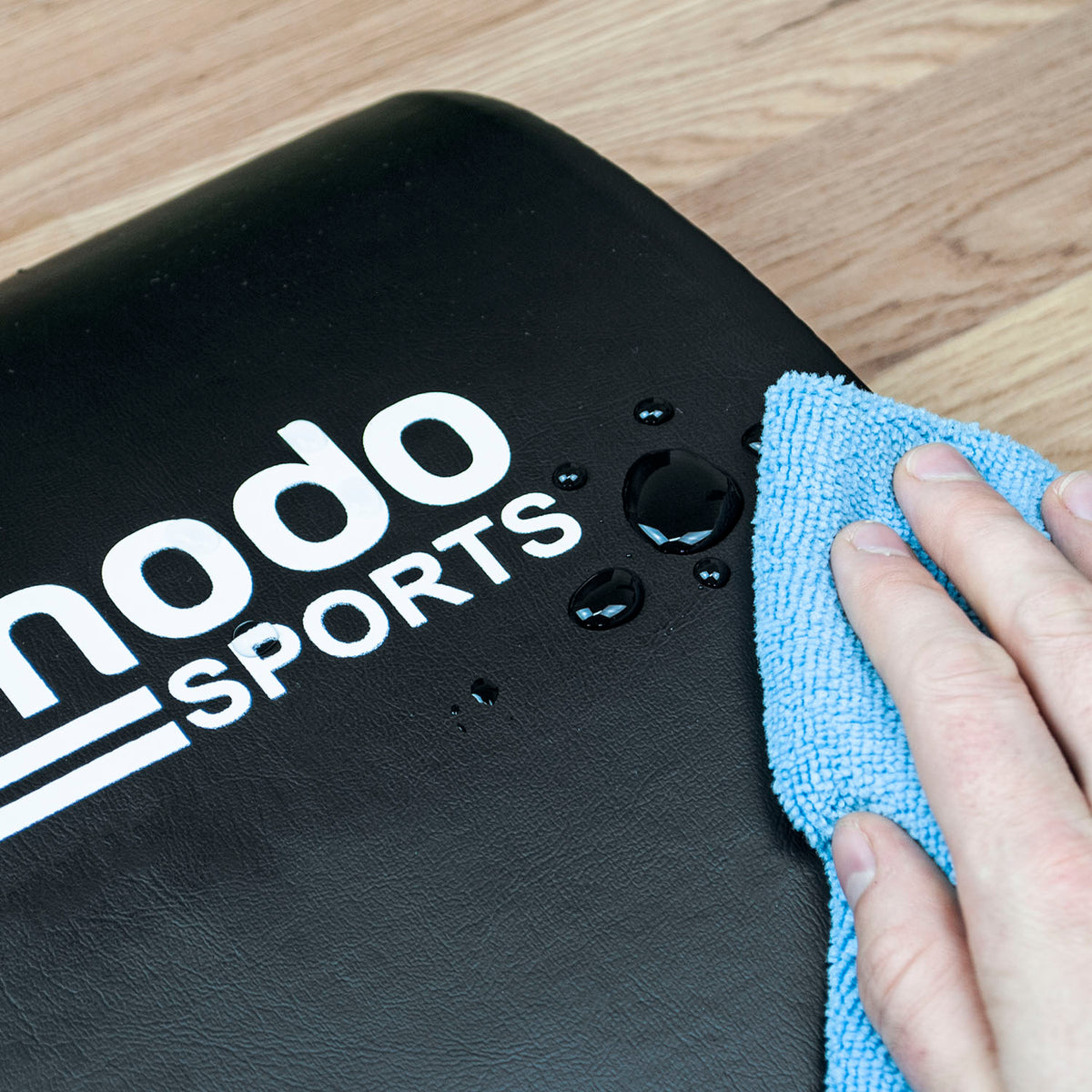 A close-up of a black Komodo Sports ab pad mat with white logo text, showing water droplets on its surface while a hand wipes it with a blue microfiber cloth on a wooden floor.
