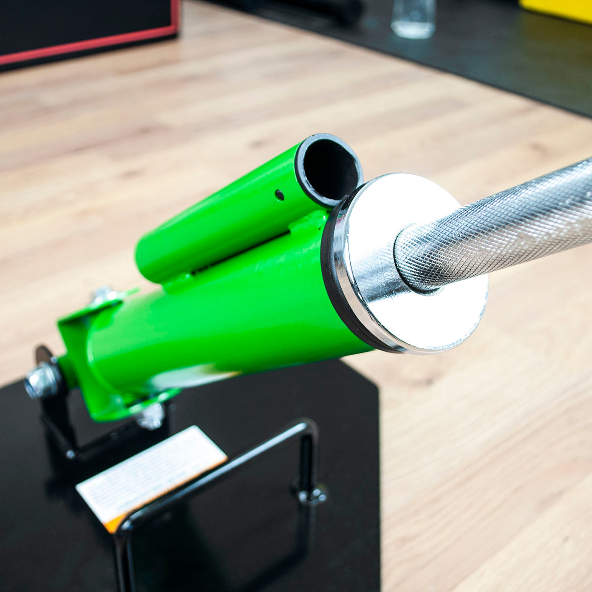 A close-up of a bright landmine row platform and silver knurled barbell rod mounted on a black display stand, set on a light wood floor.