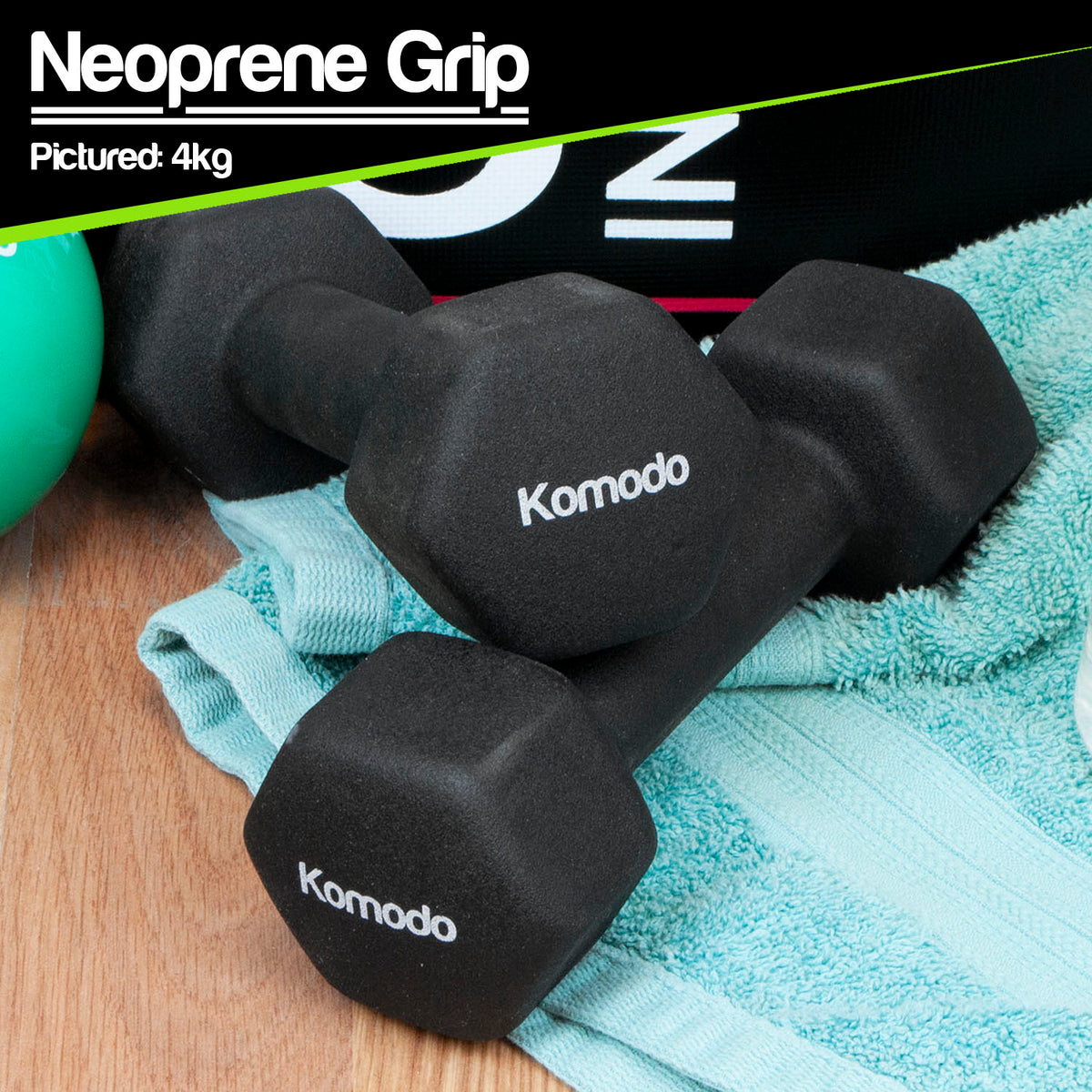 Two black Komodo neoprene hex dumbbells resting on a light blue towel over a wooden surface, with a green exercise ball partially visible and a black banner at the top reading 'Neoprene Grip Pictured: 4kg'.