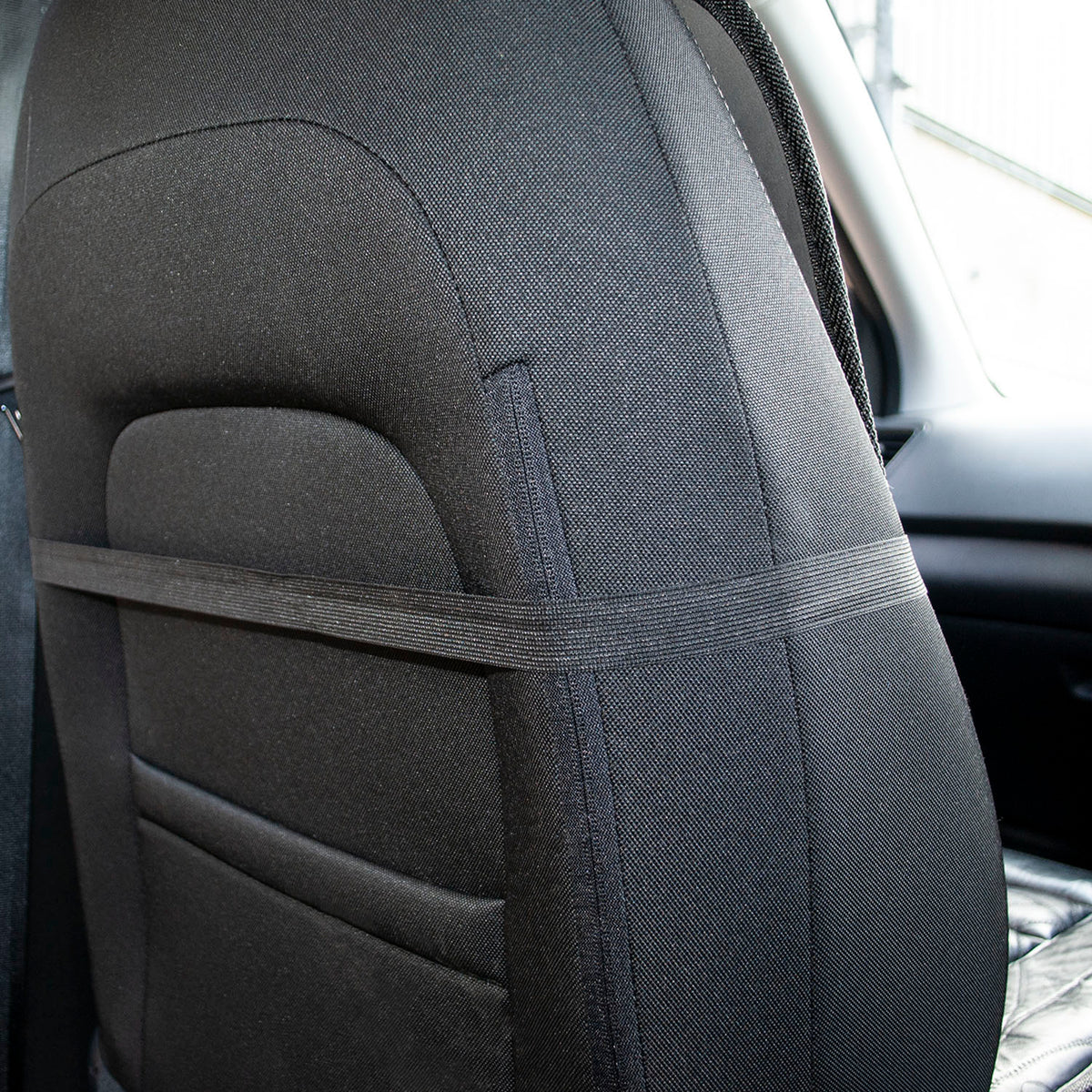 A close-up of a black car seat cover installed on a vehicle's backrest, with a horizontal support strap across the middle and a side belt loop visible.