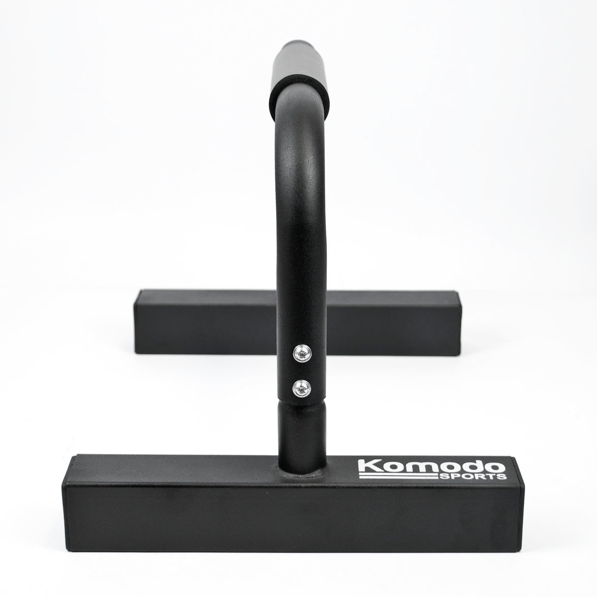 Black metal push-up bar with a wide rectangular base and a curved vertical bar, shown on a clean white background. The brand name 'Komodo Sports' is printed in white on the front of the base.