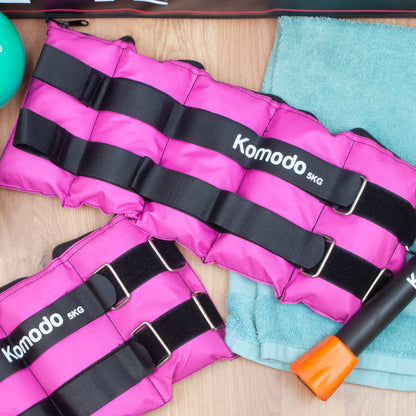 Pink Komodo 5KG ankle weights arranged on a light wood surface next to a teal towel, with a small teal ball and part of an orange-and-black barbell visible.