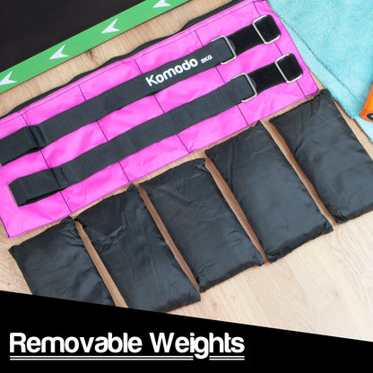 A bright pink Komodo 5kg ankle weight laid flat on a light wood surface, with several black fabric removable weight bags arranged in front. A black banner at the bottom reads 'Removable Weights.
