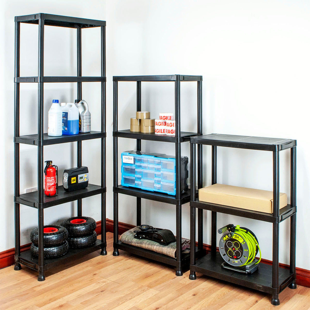 Three black plastic shelving units of different heights arranged on a wood floor against a white wall, holding assorted household and storage items.