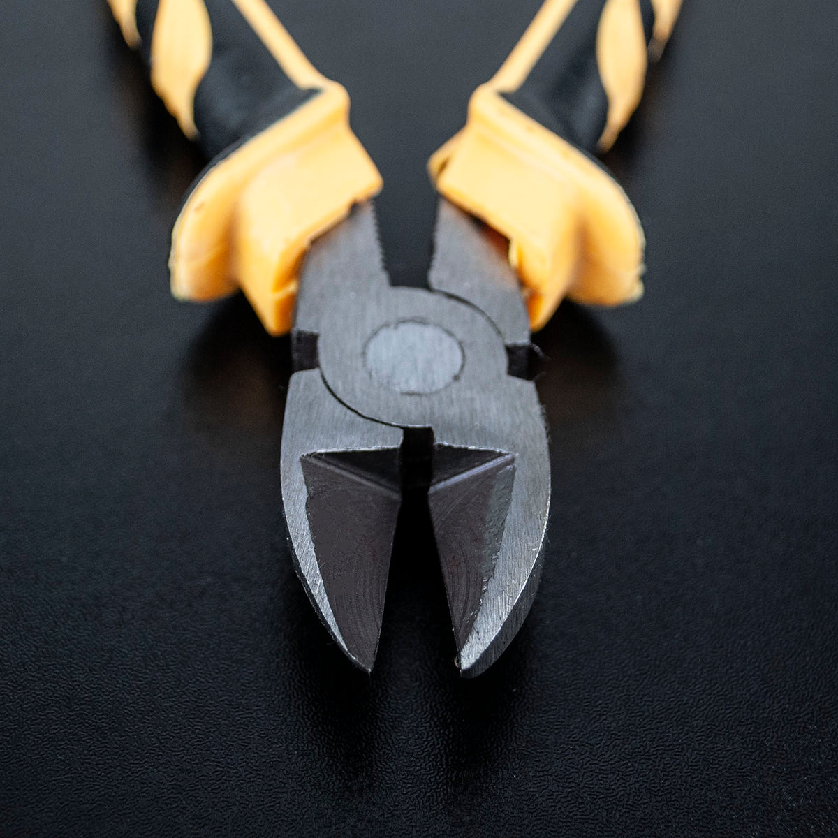 Close-up of a pair of wire cutting pliers with black and yellow striped handles and dark gray metal jaws, resting on a black textured surface.