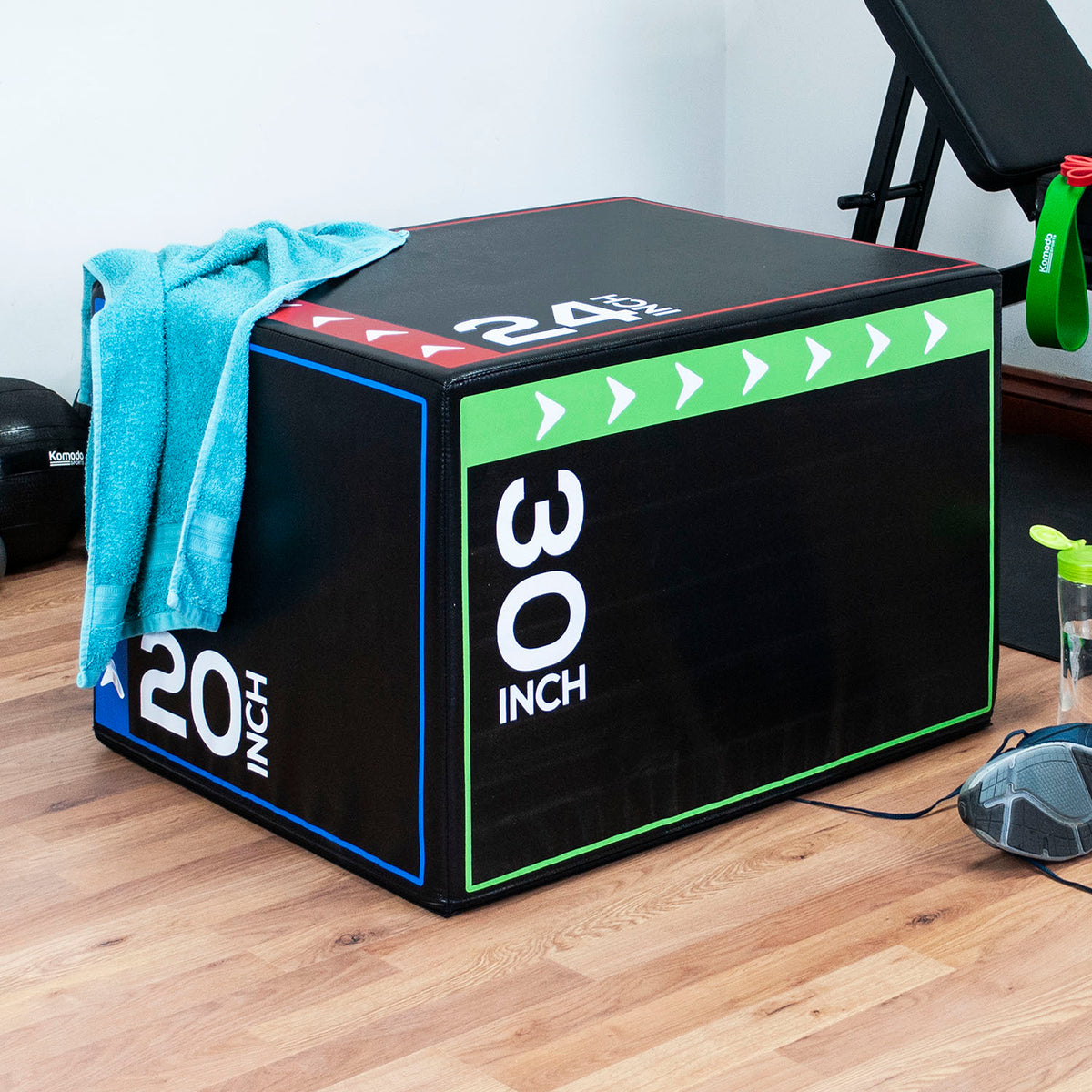 A black plyo box with green, blue, and red trim, marked 30 inch and 20 inch, sitting on a wooden floor in a gym setting with a towel draped over it.
