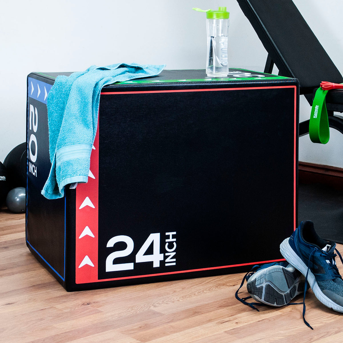 A black 24-inch plyo box with red and white markings, set on a wooden floor in a gym. A light blue towel is draped over the top, along with a clear water bottle, a green resistance band, and athletic shoes nearby.