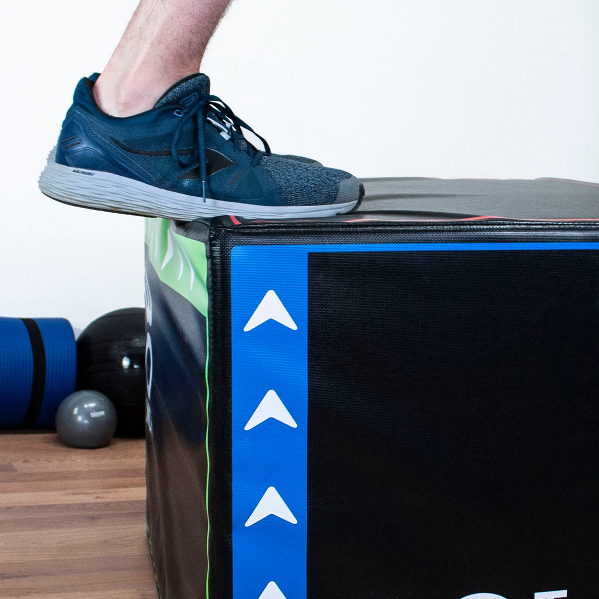 A person wearing a dark blue running shoe steps onto a black plyo box with a blue side stripe and white arrow graphics, in an indoor setting with exercise balls and a wooden floor visible.