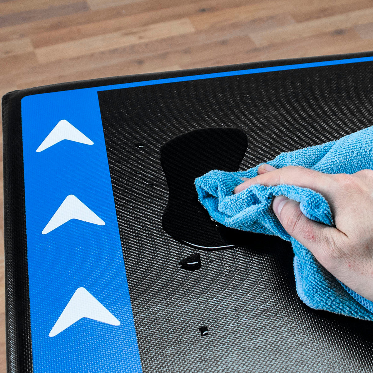 A close-up of a black jump box with a bright blue border and white arrow graphics, being wiped with a blue towel by a hand. A small puddle of liquid is visible on the mat over a wood floor background.