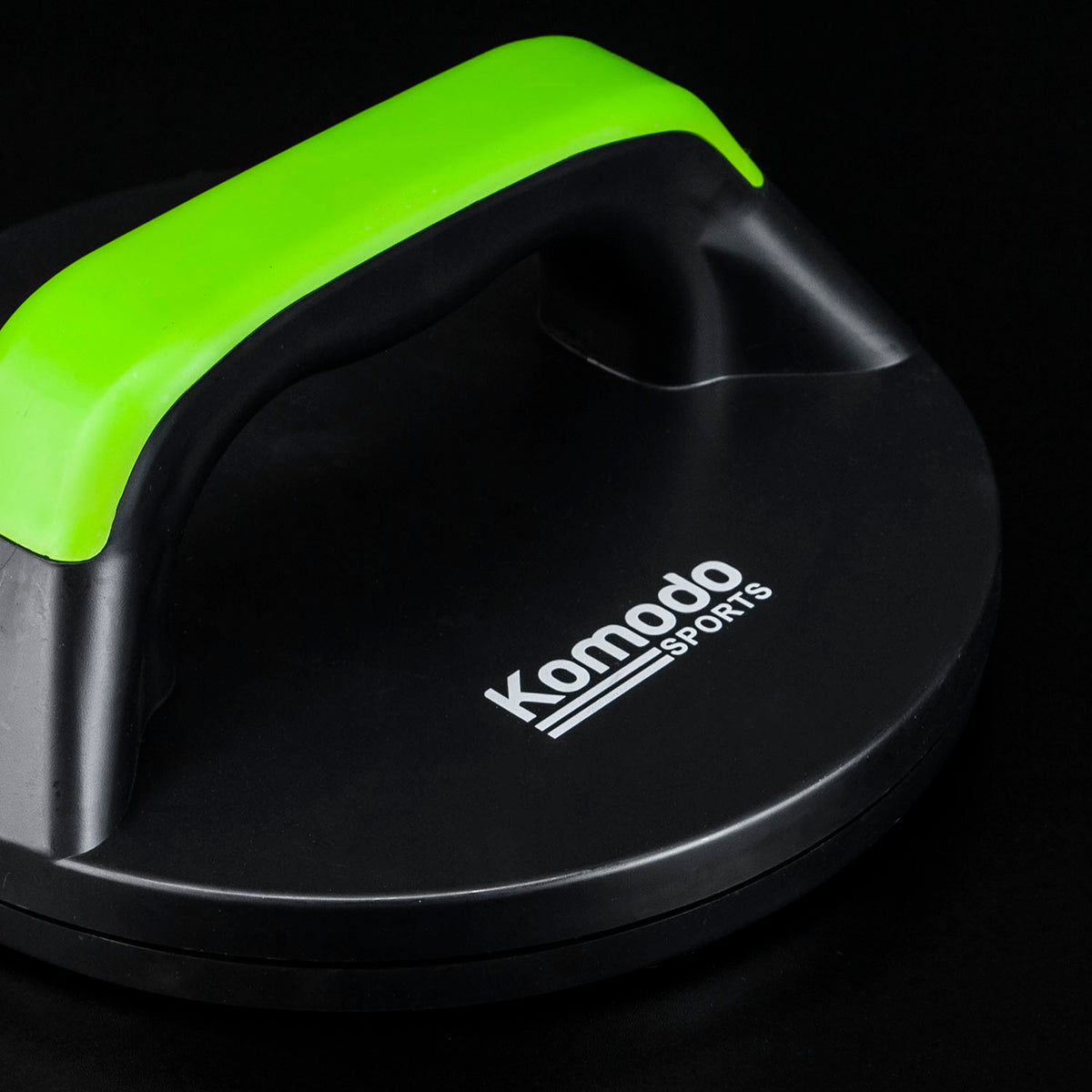 A close-up of a Komodo Sports push-up handle with a black base and a bright green handle, set against a solid black background.