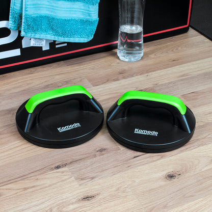 Two black Komoda push-up handles with bright green handles placed on a light wood floor, with a glass of water and a black storage box in the background.
