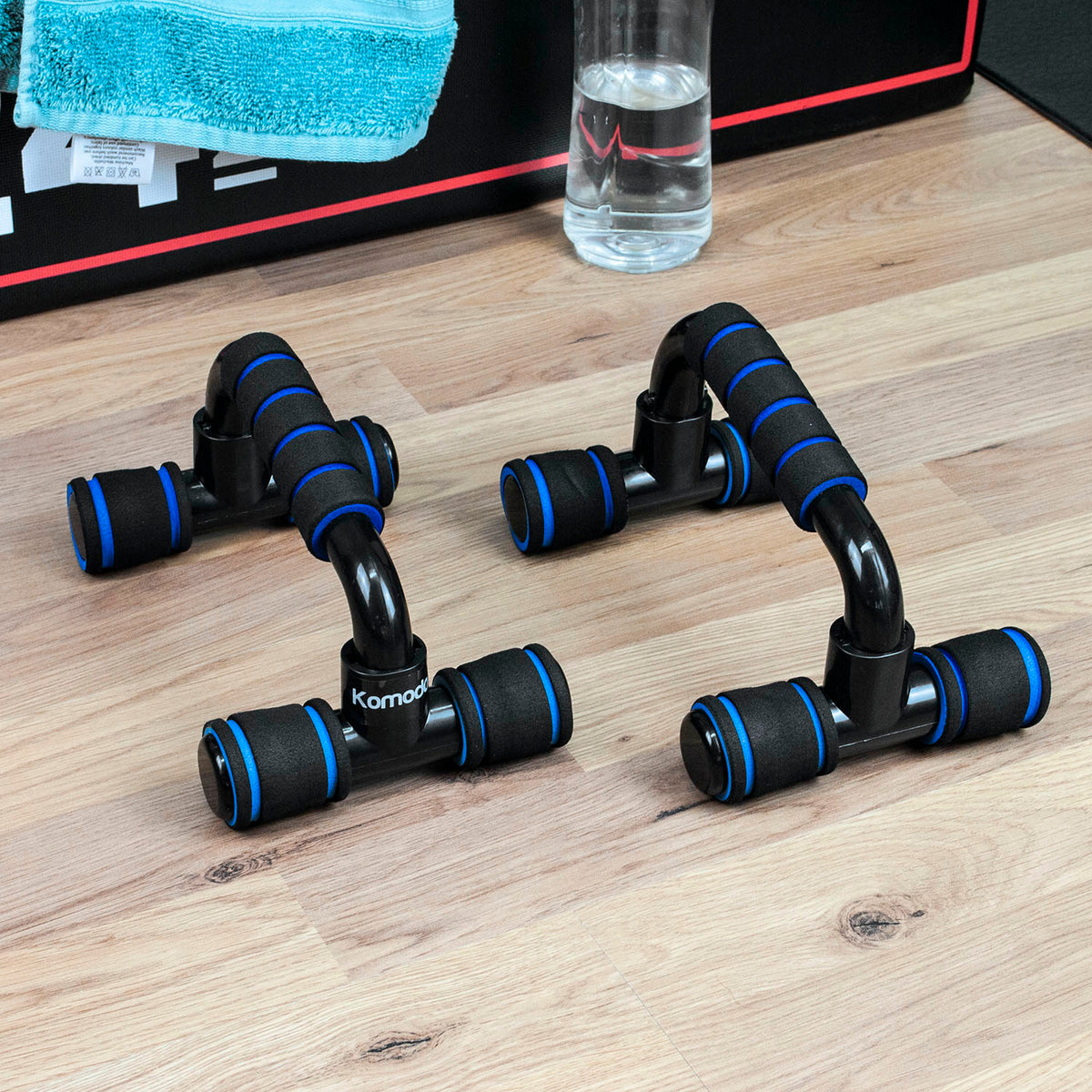 Two black push-up support bars with blue accent rings, displayed on a light wood floor next to a towel and a glass of water.