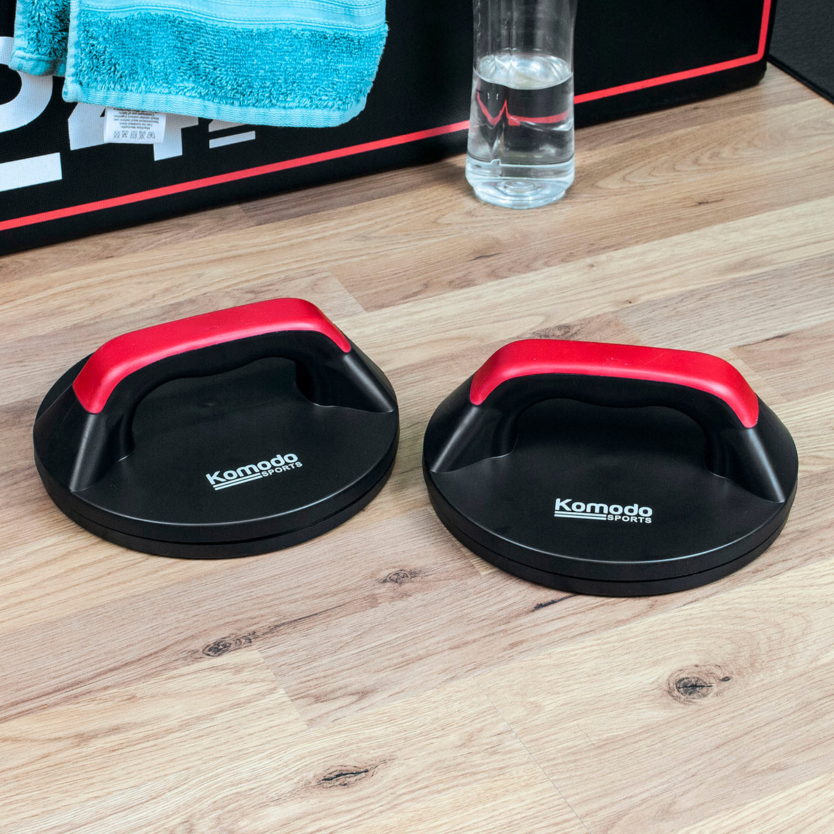 Two black Komodo Push Ups with red handles placed on a light wood floor, with a towel and water bottle in the background.