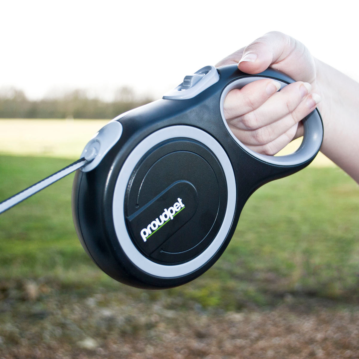 A hand holding a black and gray Proudpet retractable dog leash with a metal leash rod extended, set against a blurred outdoor background of grass and trees.