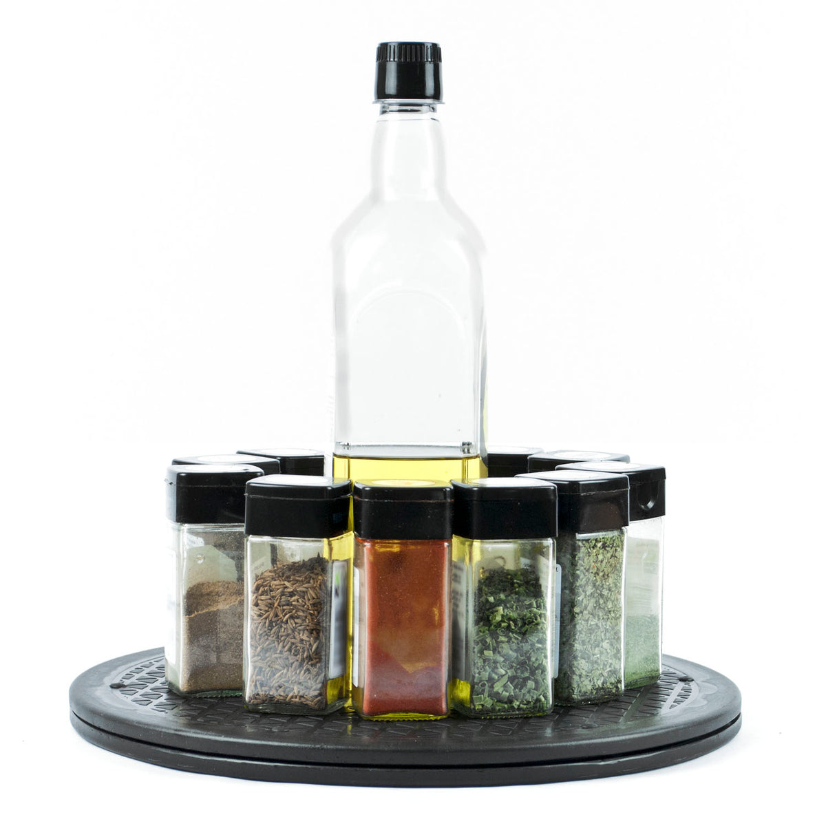 A clear glass bottle of oil stands upright in the center of a circular black tray, surrounded by eight small clear spice or herb jars with black lids, all arranged on a white background.